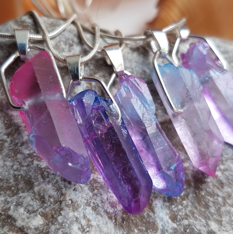 May include: Close-up of five crystal pendant necklaces. Each pendant features a raw, elongated crystal in shades of pink, purple, and blue, set in a silver-tone metal frame. The necklaces are displayed on a textured grey surface.