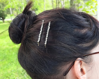 May include: Two silver-toned hair pins featuring a single row of clear, round-cut rhinestones. The hair pins are secured into dark brown hair styled in a bun, positioned side-by-side to hold the hair in place against the scalp.