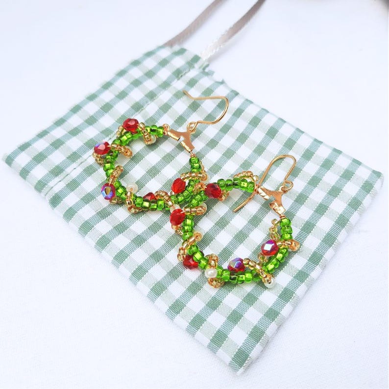 May include: Handmade hoop earrings with a festive wreath design. The earrings have a gold-tone metal base, decorated with green and red beads. They are displayed on a green and white gingham fabric, evoking a seasonal theme.