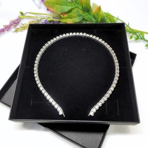 May include: A silver headband with clear rhinestones set in a square shape. The headband is in a black box.