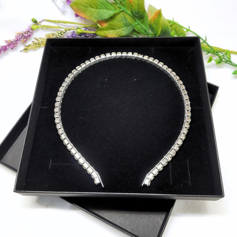 May include: A silver headband with clear rhinestones set in a square shape. The headband is in a black box.