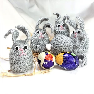 May include: Five gray crocheted bunny rabbits with large black eyes and pink noses, sitting on a bed of straw with two foil-wrapped chocolate Easter eggs.