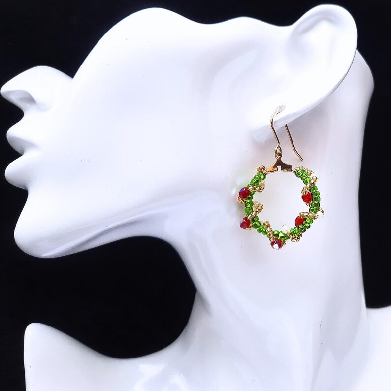 May include: Gold-coloured hoop earrings with a wreath design. The wreaths are decorated with green and red beads, creating a festive appearance. The earrings are displayed on a white mannequin head against a black background.