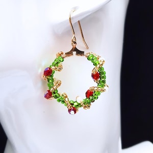May include: A festive earring featuring a beaded wreath design. The wreath is crafted from green beads, accented with red and white beads, and gold-coloured detailing. The earring hangs from a gold-coloured hook.