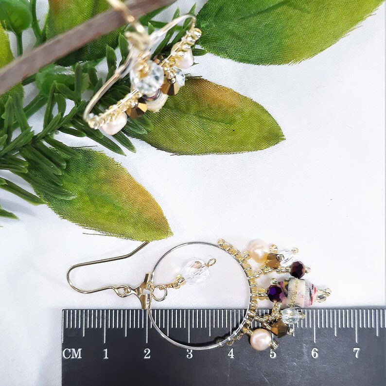 Pu&ograve; includere: Orecchini a cerchio dorati con un ciondolo pendente che presenta un mix di perline trasparenti, viola e rosa, e dettagli dorati. Gli orecchini sono misurati con un righello, mostrando che sono lunghi circa 6 cm.