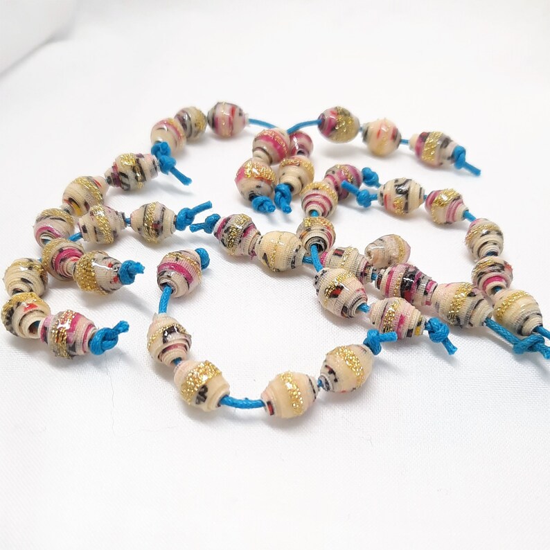 Pu&ograve; includere: Una collana di perline di carta colorate con un motivo bianco, rosa e oro. Le perline sono collegate da uno spago turchese.
