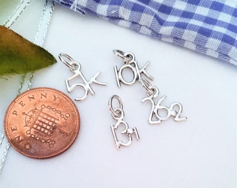 May include: Three silver-toned metal charms shaped as the numbers 5K, 10K, and 13.1, alongside a 26.2 charm. A copper-coloured one penny coin is placed next to the charms for scale, showing the relative size of the small jewellery pieces.
