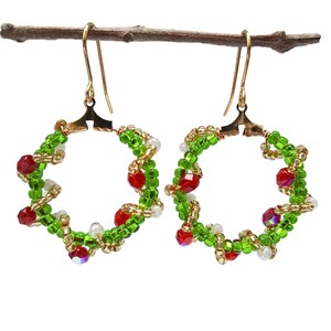 May include: A pair of festive earrings with a wreath design. The earrings are made with green, red, and white beads, and gold-coloured wire. The earrings hang from gold-coloured ear wires, ideal for Christmas or other celebrations.