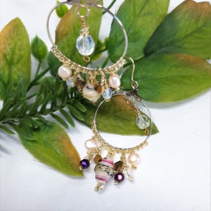 Pu&ograve; includere: Orecchini a cerchio dorati con un ciondolo pendente decorato con una variet&agrave; di perline, tra cui perle bianche, cristalli trasparenti e una piccola perlina rivestita in tessuto con accenti rosa e viola.