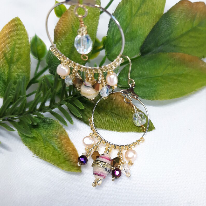 Pu&ograve; includere: Orecchini a cerchio dorati con un ciondolo pendente decorato con una variet&agrave; di perline, tra cui perle bianche, cristalli trasparenti e una piccola perlina rivestita in tessuto con accenti rosa e viola.