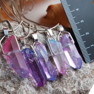 May include: Five crystal pendant necklaces with silver-coloured metal findings. The crystals are various colours, including pink, purple, and blue. A ruler is in the background, with the crystals measuring approximately 2.5 to 5 cm in length. The necklaces are on a grey stone surface.