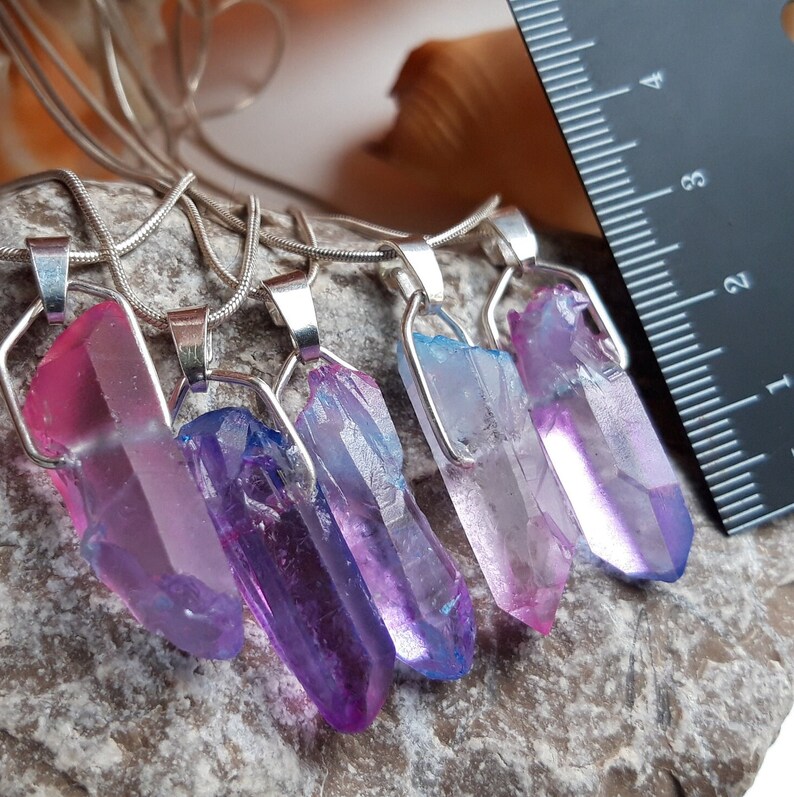 May include: Five crystal pendant necklaces with silver-coloured metal findings. The crystals are various colours, including pink, purple, and blue. A ruler is in the background, with the crystals measuring approximately 2.5 to 5 cm in length. The necklaces are on a grey stone surface.