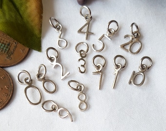 May include: A collection of silver-toned metal number charms, each featuring a small circular jump ring at the top. The set includes individual digits and the number 20, all displayed with a metallic finish on a plain white surface.