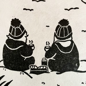 Linoprint of two wild swimmers enjoying pastries on the beach. Close up of wild swimmers. Printed in black ink with a red sun.