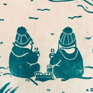 Linoprint of two wild swimmers enjoying pastries on the beach. Close up of the wild swimmers. Printed in turquoise ink with a red sun.