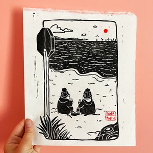 Linoprint of two wild swimmers enjoying pastries on the beach. Printed in black ink with a red sun.