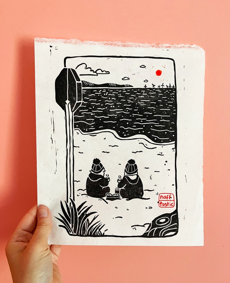 Linoprint of two wild swimmers enjoying pastries on the beach. Printed in black ink with a red sun.