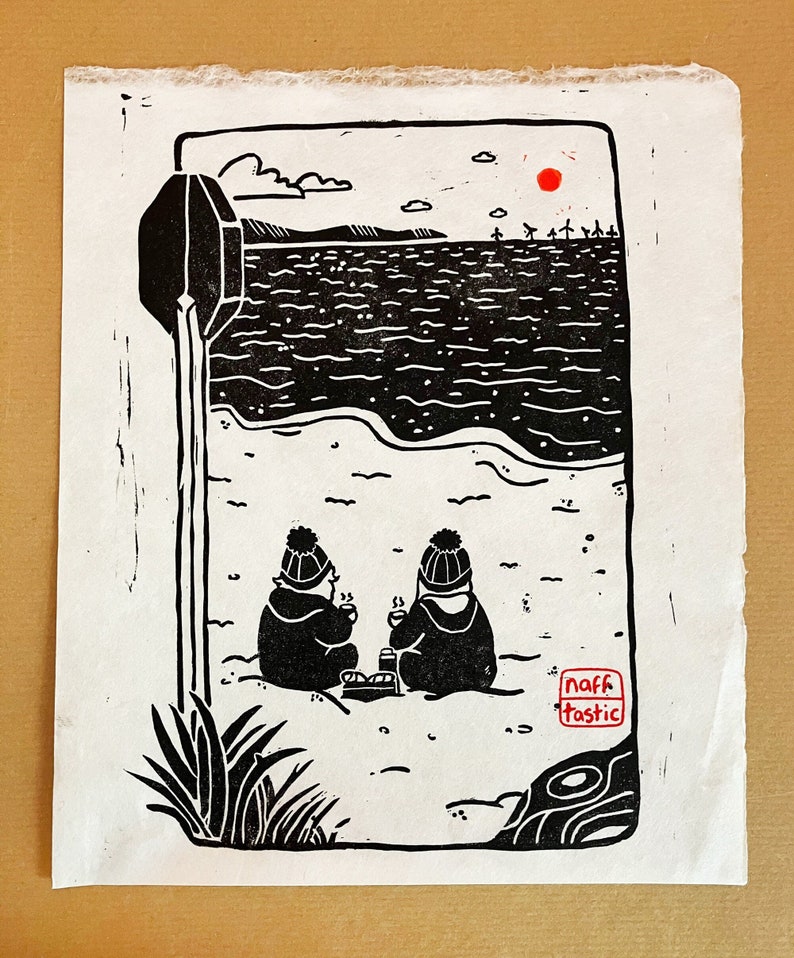 Linoprint of two wild swimmers enjoying pastries on the beach. Printed in black ink with a red sun.