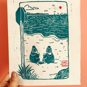 Linoprint of two wild swimmers enjoying pastries on the beach. Printed in turquoise ink with a red sun.