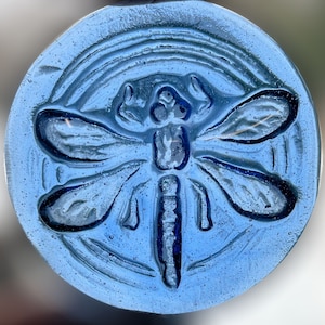 May include: A round, light blue glass piece with a dragonfly design. The dragonfly is centered, with detailed wings and body. The design is embossed, creating a textured surface. The piece has a subtle, iridescent quality.