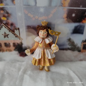 May include: A handcrafted doll dressed in a gold and white costume, holding a gold mask on a stick and a white rose. The doll has a gold crown and is set against a winter-themed backdrop.