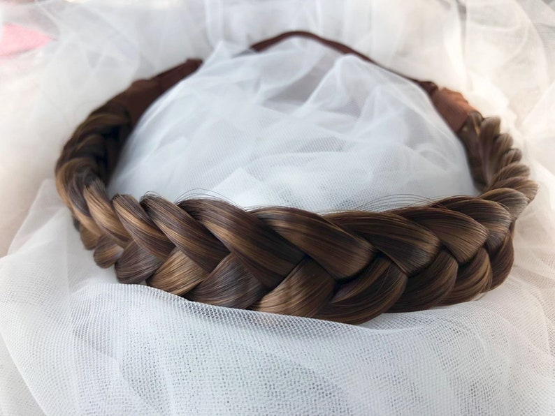 May include: A brown braided hairpiece with a brown elastic band. The hairpiece is on a white background.
