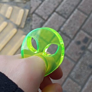 May include: A neon green alien-shaped ring. The ring has two oval eye cutouts and a circular design. The ring is held up by a finger. The background is a brick sidewalk with yellow tactile paving.