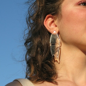 May include: A woman with curly brown hair wearing a white vest top and large, clear, geometric earrings. She is looking up and smiling.