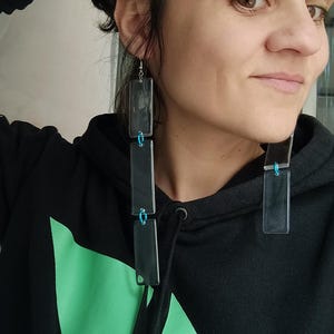 May include: Long, rectangular earrings with a dark, translucent appearance. Each earring is composed of three connected sections, linked by small blue accents. The earrings are worn with a black hoodie featuring a green geometric design.