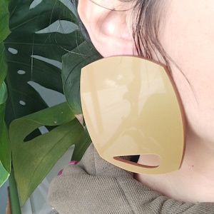 Könnte beinhalten: Ein großer, hellbrauner, geometrisch geformter Ohrring mit einem Ausschnitt. Der Ohrring ist aus einem glatten, glänzenden Material gefertigt.