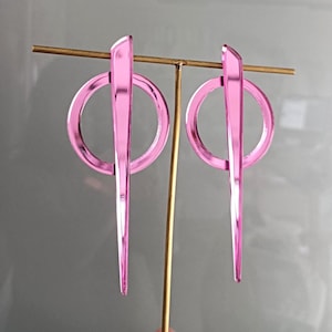 May include: A pair of pink acrylic earrings. Each earring features a circular shape with a vertical, elongated, pointed element. The earrings are displayed on a gold-colored stand against a gray background. The earrings have a modern, geometric design.