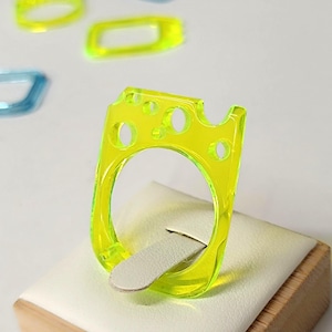 May include: A neon yellow ring with a rectangular design and circular cutouts. The ring is displayed on a white cushion atop a wooden block. Other rings in blue and yellow are in the background.