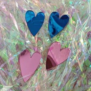 May include: Three heart-shaped decorative pieces, two are blue and one is pink. The hearts are made of reflective material and are arranged on a iridescent background.