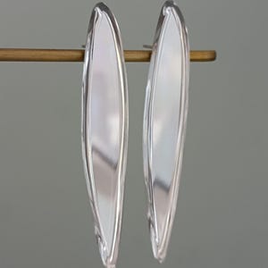 Puede incluir: Un par de pendientes plateados, alargados con forma de hoja. Los pendientes se muestran sobre una barra dorada sobre un fondo gris. Los pendientes tienen una superficie lisa y reflectante.