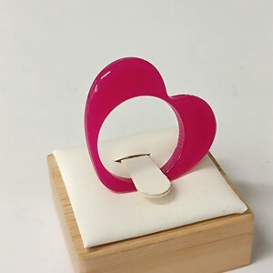 May include: A vibrant magenta heart-shaped ring. The ring is displayed on a white cushion, set on a small wooden block. The ring's design is a solid, continuous heart shape.