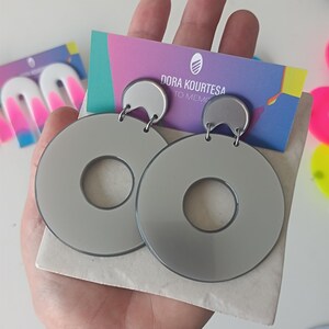 Könnte beinhalten: Ein Paar großer, grauer, kreisförmiger Ohrringe mit einem kleineren, silbernen, halbmondförmigen Oberteil. Die Ohrringe werden in einer Hand gehalten, mit einer Visitenkarte mit der Aufschrift "Dora Kourtesa To Memo". Im Hintergrund sind weitere farbenfrohe Ohrringe zu sehen.