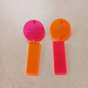 Könnte beinhalten: Ein Paar leuchtende Neon-Ohrringe. Jeder Ohrring hat einen kreisförmigen oberen Teil und einen rechteckigen unteren Teil, verbunden durch eine kleine silberne Schlaufe. Ein Ohrring ist rosa mit einem orangefarbenen Unterteil, der andere orange mit einem rosa Unterteil. Die Ohrringe sind aus durchscheinendem Material.