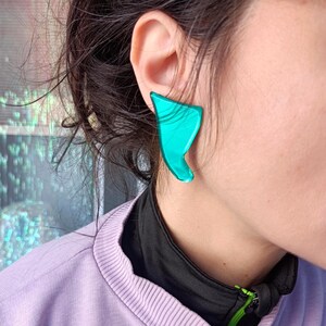 Puede incluir: Primer plano de una persona que lleva un par de pendientes de forma abstracta de color turquesa. Los pendientes tienen un acabado brillante y están colocados en la oreja. La persona lleva una chaqueta de color lila claro con cuello negro.