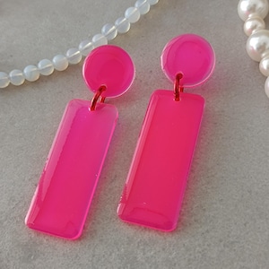 Op de afbeelding: Een paar roze acryl oorbellen met een ronde bovenkant en een rechthoekige onderkant. De oorbellen zijn in een heldere, neon roze kleur.