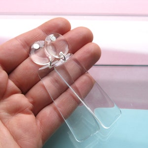 Peut inclure: Une paire de boucles d'oreilles en acrylique transparent avec une forme rectangulaire et un dessus rond. Les boucles d'oreilles sont pendantes et ont un crochet argenté.