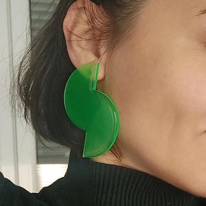 Puede incluir: Un par de pendientes grandes de acrílico verde brillante. Los pendientes tienen una forma única y abstracta, con un diseño curvo y semicircular. Los pendientes son de un color verde translúcido vibrante.