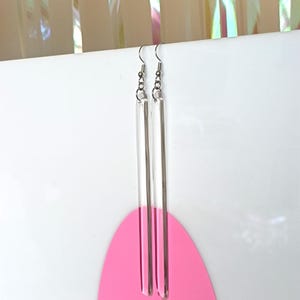 Puede incluir: Un par de pendientes largos de acrílico transparente con ganchos plateados. La parte inferior de cada pendiente está sumergida en rosa.