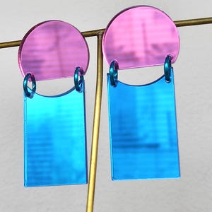 May include: A pair of modern earrings with a geometric design. Each earring features a pink, semi-circular top and a blue, rectangular bottom. The two pieces are connected by a small, teal-colored ring. The earrings are displayed on a gold-colored stand.