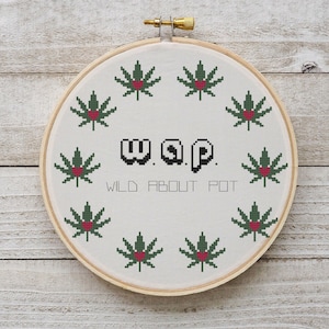 May include: A white embroidery hoop with a green and red cross-stitch design of marijuana leaves with hearts in the center. The text "w.a.p. WILD ABOUT POT" is stitched in black.