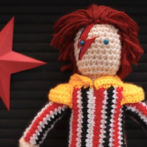 May include: Crocheted doll with red hair, a yellow, black and white striped outfit, and a red lightning bolt painted on its face. The doll is standing in front of a black background with the words "ZIGGY STARDUST" written below it.