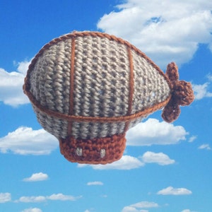 以下が含まれることがあります： グレーとブラウンの毛糸で編まれた飛行船。ブラウンのプロペラが付いている。飛行船は青い空と白い雲を背景にしている。