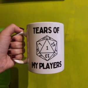 Può includere: Tazza in ceramica bianca con testo nero che recita "Tears of My Players" e un'illustrazione in bianco e nero di un dado a venti facce con il numero uno tirato.
