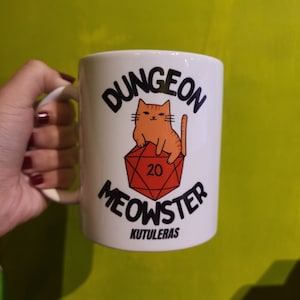 Peut inclure: Une tasse en céramique blanche avec un dé rouge à 20 faces, un chat orange dessiné assis dessus et le texte "Dungeon Meowster" en noir. Le texte "Kutuleras" est imprimé en noir sous le dé.