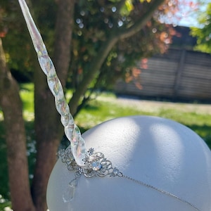 Puede incluir: Una diadema de unicornio plateada con un cuerno transparente iridiscente y detalles de cristal. La diadema está en una cabeza de maniquí blanca.