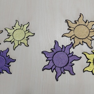 Peut inclure: Écussons brodés en forme de soleil de différentes couleurs, dont or, jaune et violet, avec des contours noirs. Les soleils ont un aspect texturé et détaillé, disposés sur une surface claire.